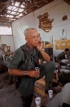 an older man sitting on a bench talking on a cell phone in a room full of other people