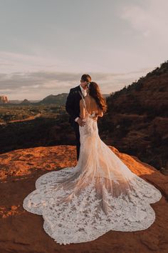 a bride and groom standing on top of a mountain in their wedding dress with the sun setting behind them