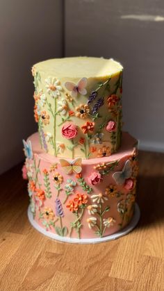 two tiered cake decorated with flowers and butterflies on a wooden countertop, in front of a white wall