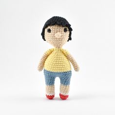 a small crocheted doll with black hair and blue pants, standing in front of a white background