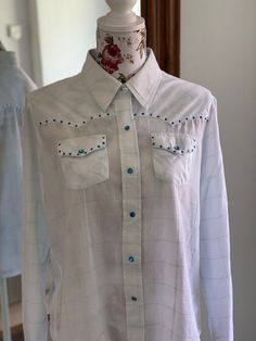 "White country and western blouse with a check pattern with a small silver stripe. The blouse is adorned with blue plastic stones. The blouse is closed with pushbuttons with clear blue plastic stones as well. The cuffs is closed with pushbuttons as well. The fabric is 100% cotton. The blouse is tagged as size XL but please check measures below. Measured with blouse laid flat and buttons closed. Armpit to armpit: 62 cm - 24,4\" Waist: 55 cm - 21,7\" Lenght: 66 cm - 26\"" Western Style Blue Tops With Buttons, Western Blue Button-up Top, Western Long Sleeve Blouse For Rodeo, Blue Western Tops For Rodeo, Blue Tops With Snap Buttons For Rodeo, Blue Rodeo Top With Snap Buttons, Blue Snap Button Tops For Rodeo, Blue Snap Button Top For Rodeo, Western Style Buttoned Tops For Rodeo