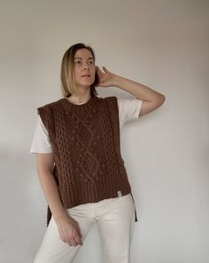 a woman standing in front of a white wall talking on a cell phone and wearing a brown knitted vest