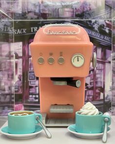 an orange and blue coffee maker sitting on top of a table next to two cups