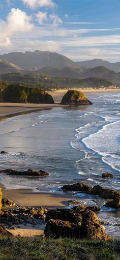 an ocean view with mountains in the distance and waves crashing on the beach, along with green grass