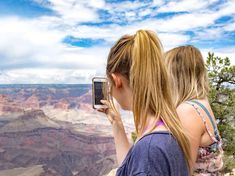 two girls looking at the grand canyon with a cell phone in their hands while standing on a cliff