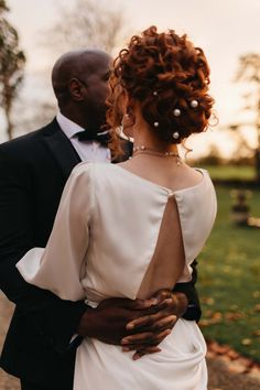 Groom in black tux with bowtie embracing bride in open back detail satin wedding dress with pearl hair accessories and pearl necklace Curly Updo With Pearls, Pearl Curly Hairstyles, Boho Chic Hairstyles Wedding, Curly Hair Pearls, Curly Updos For Wedding, Bridal Updo Pearls, Pearl Wedding Hairstyles, Shoulder Length Curly Wedding Hair, Pearl Pins In Hair