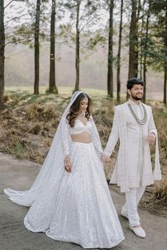 a bride and groom in white outfits holding hands while walking through the woods with trees behind them