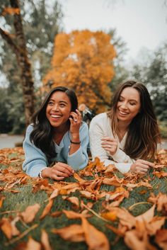 two women laying in the leaves talking on their cell phones and smiling at the camera