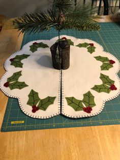 a christmas tree skirt with holly leaves and red berries is on a cutting board next to a small pine tree