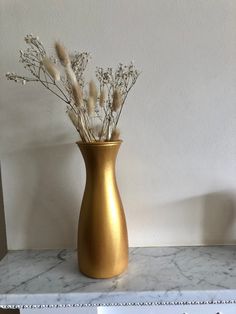 a gold vase filled with dried flowers on top of a marble counter next to a white wall