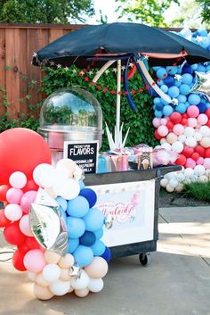 an umbrella and balloons are set up in front of a cake cart for a birthday party