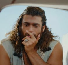 a man with long hair wearing a vest and holding his hand to his face while sitting in front of a mirror