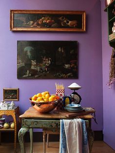 a bowl of fruit is sitting on a table in front of two paintings and a lamp