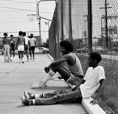 two young men sitting on the side of a road next to a fence and basketball hoop