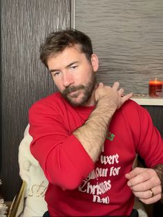 a man in a red shirt sitting down with his hands on his chest and looking at the camera