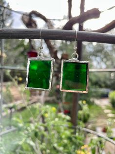 two square shaped green glass earrings hanging from a metal bar in front of some plants