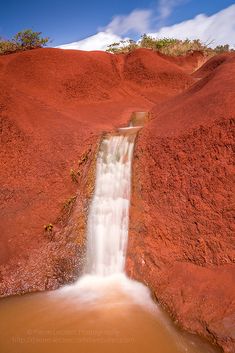 a small waterfall running down the side of a red hill next to a body of water