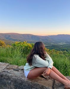 a woman sitting on top of a large rock next to a lush green field and mountains