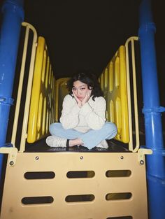 a young woman sitting on top of a yellow and blue playground slide with her hands under her chin