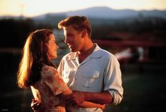 a man and woman standing next to each other in front of a field at sunset