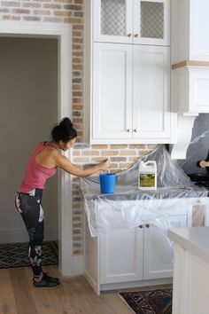 a woman is painting a kitchen wall with white cabinets and she has a blue bucket in her hand