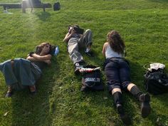 three people laying on the grass with their backs to each other and backpacks behind them