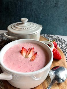 a bowl of strawberry smoothie on a wooden board next to a spoon and pot