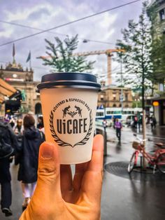 a hand holding up a coffee cup in the middle of a busy street with people walking around