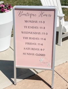 a pink sign sitting on the side of a sidewalk next to a white chair and flower pot