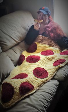 a woman is laying on a couch with a crocheted blanket and eating pizza