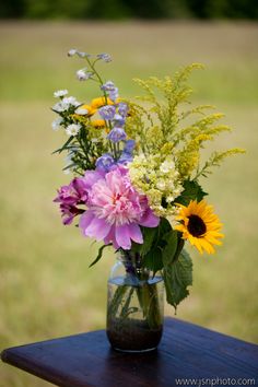 a vase filled with lots of flowers on top of a wooden table next to a field