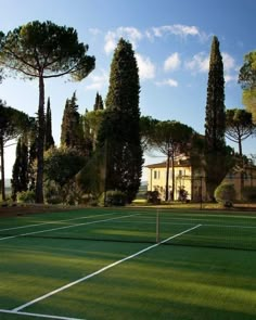 a tennis court surrounded by tall trees and grass with a house in the back ground