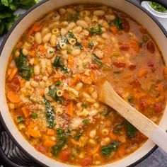 a pot filled with pasta and vegetables on top of a table next to a wooden spoon