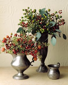 two silver vases with berries and greenery in them