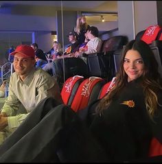 two people are sitting in the stands at an indoor sporting event, one is wearing a red hat and the other wears a black shirt