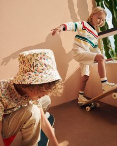 two children are playing on their skateboards and one is wearing a sun hat while the other watches