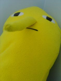 a yellow stuffed animal with eyes and nose sitting next to a white wall in a corner