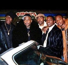 several men standing next to a car in front of a pop - n - c's sign