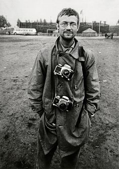 black and white photograph of man in trench coat with camera strapped to his back, standing on field