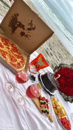 pizza, wine glasses and snacks are on the table by the beach in front of an open box