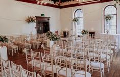 rows of white chairs with flower arrangements on them in a large room filled with windows