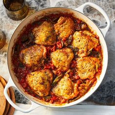 a pan filled with chicken and tomato sauce on top of a wooden cutting board next to wine glasses