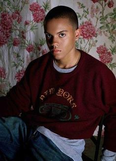 a young man sitting in front of a floral wallpaper wearing a red sweater and jeans