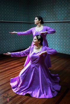 two women in purple dresses are dancing on a wooden floor with their arms stretched out