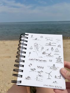 someone holding up a notebook with drawings on it in front of the ocean and beach