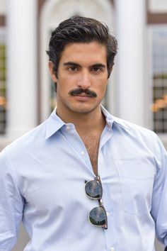 a man with a moustache wearing sunglasses and a blue shirt is standing in front of a building