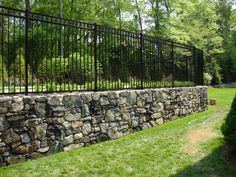 a stone wall with black iron fence in front of trees and grass on the other side