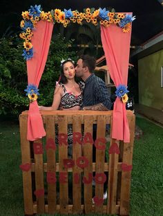 a man and woman sitting on top of a wooden box with flowers around it, in the grass