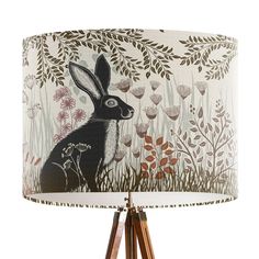 a lamp that has a rabbit on it and flowers in the background, with a wooden tripod base