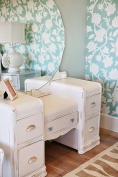 a white desk with drawers and a mirror on top of it in front of a floral wallpaper